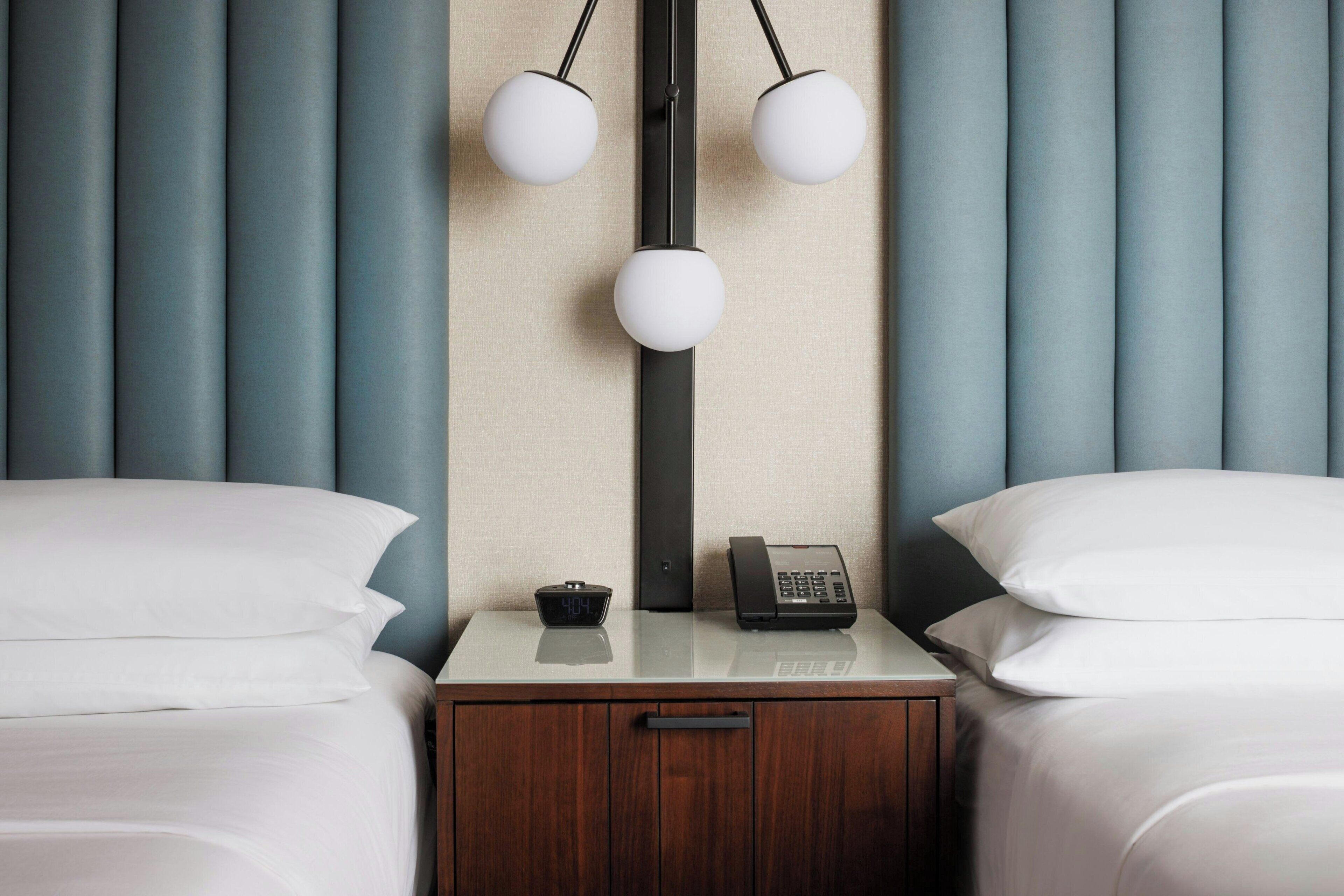 A hotel room with two beds, a bedside table with a phone and a digital clock, and a wall-mounted lamp.