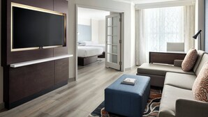 A modern hotel room with a flat-screen TV, a sofa, a blue ottoman, a bed, and a large window with sheer curtains. - Chicago Marriott Suites Deerfield (Deerfield)
