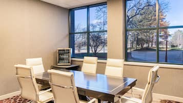 Un salón de conferencias con una gran mesa y seis sillas, ventanas amplias que dan a vistas de árboles y un estacionamiento.