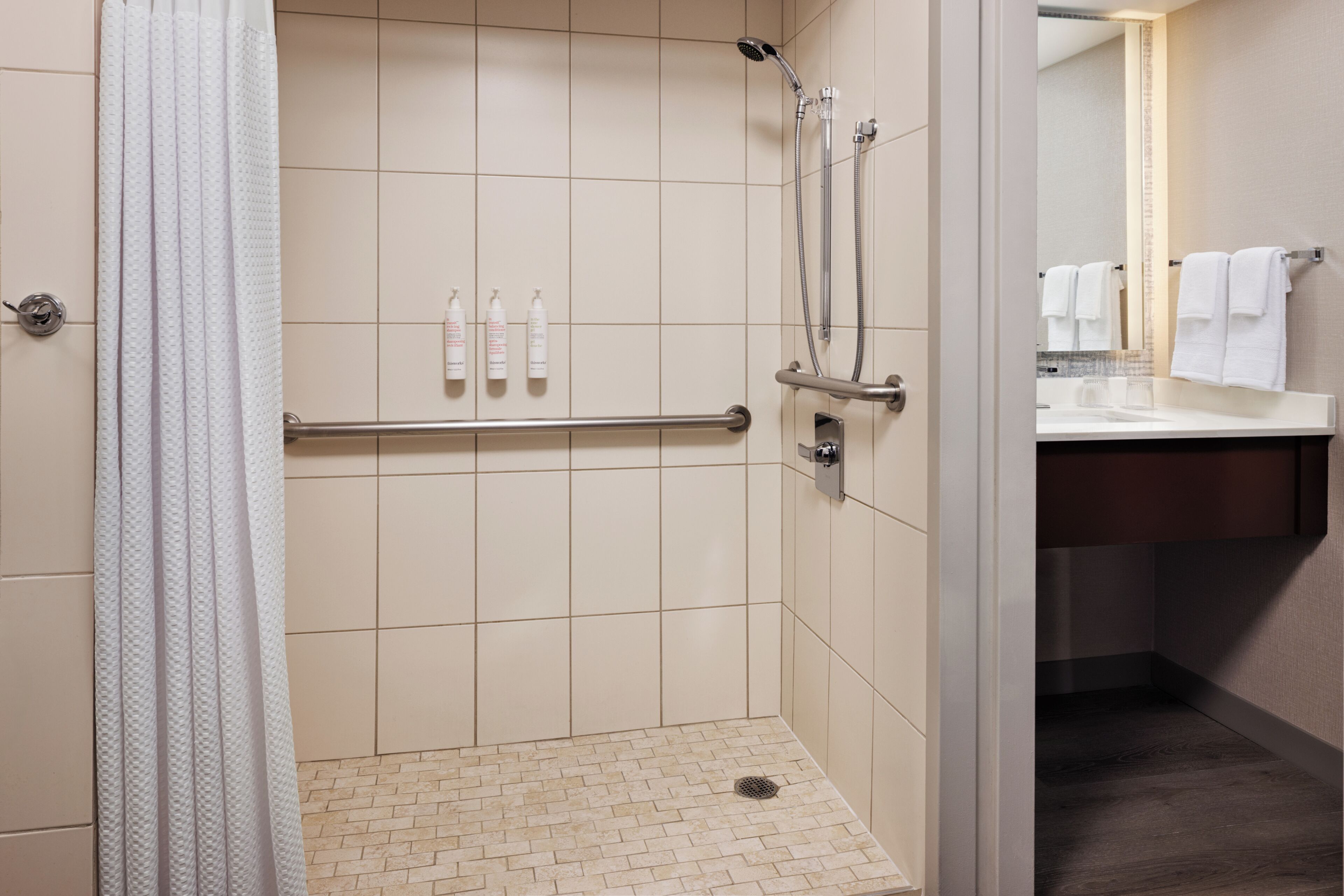 A shower with a handrail, a tiled floor, and a bathroom with a sink and towel rack.
