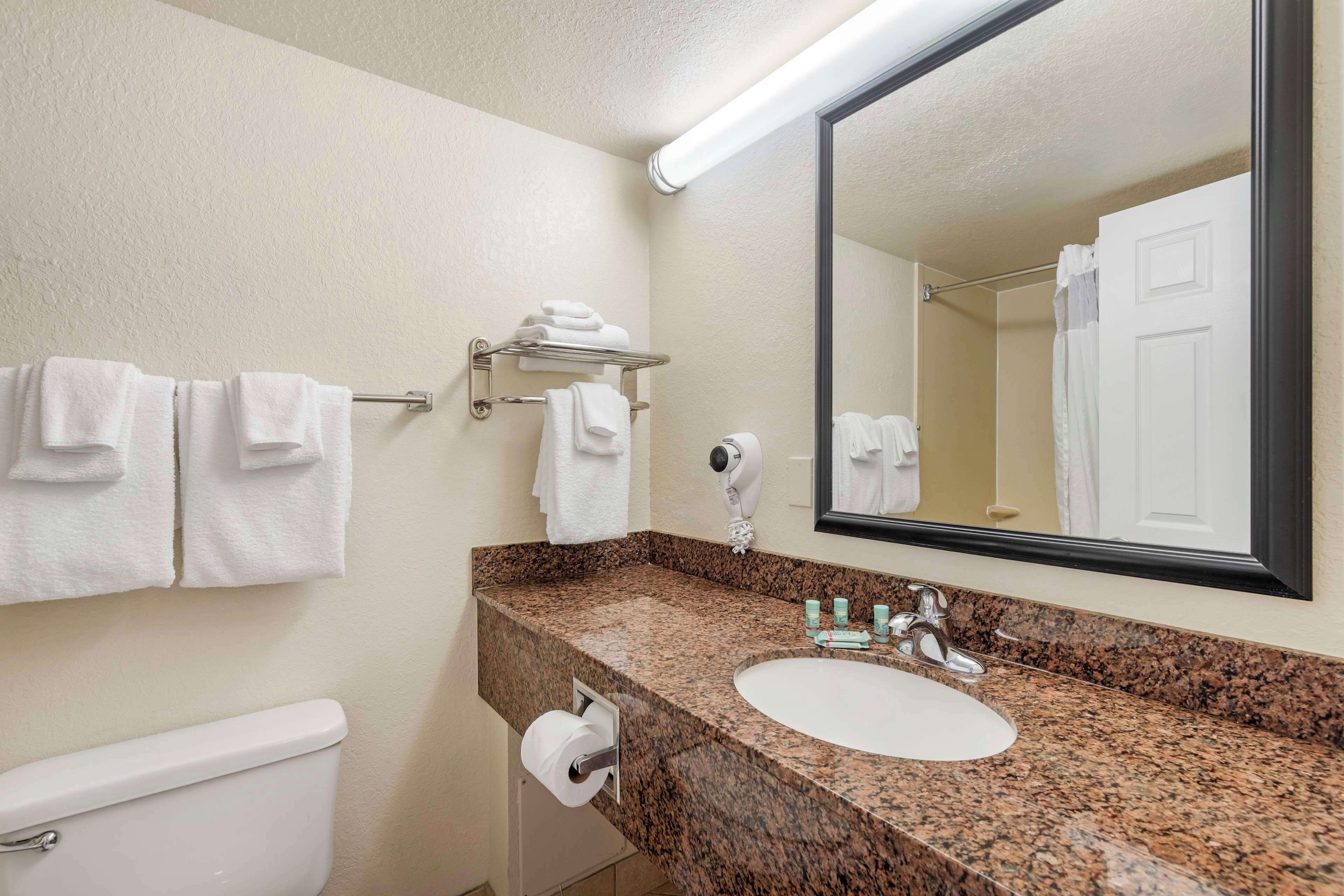 A bathroom with a white toilet, a sink with a granite countertop, a towel rack with folded towels, a hand dryer, and a large mirror.