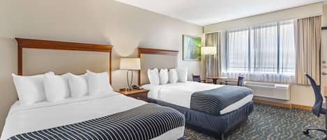 A hotel room with two beds, a desk, a chair, and a large window with curtains.