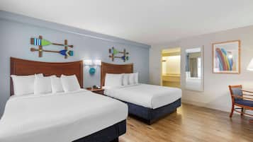 Quarto de hotel com duas camas, cabeceiras de madeira, roupa de cama branca, piso de madeira, uma pequena mesa com uma cadeira e arte decorativa na parede.
