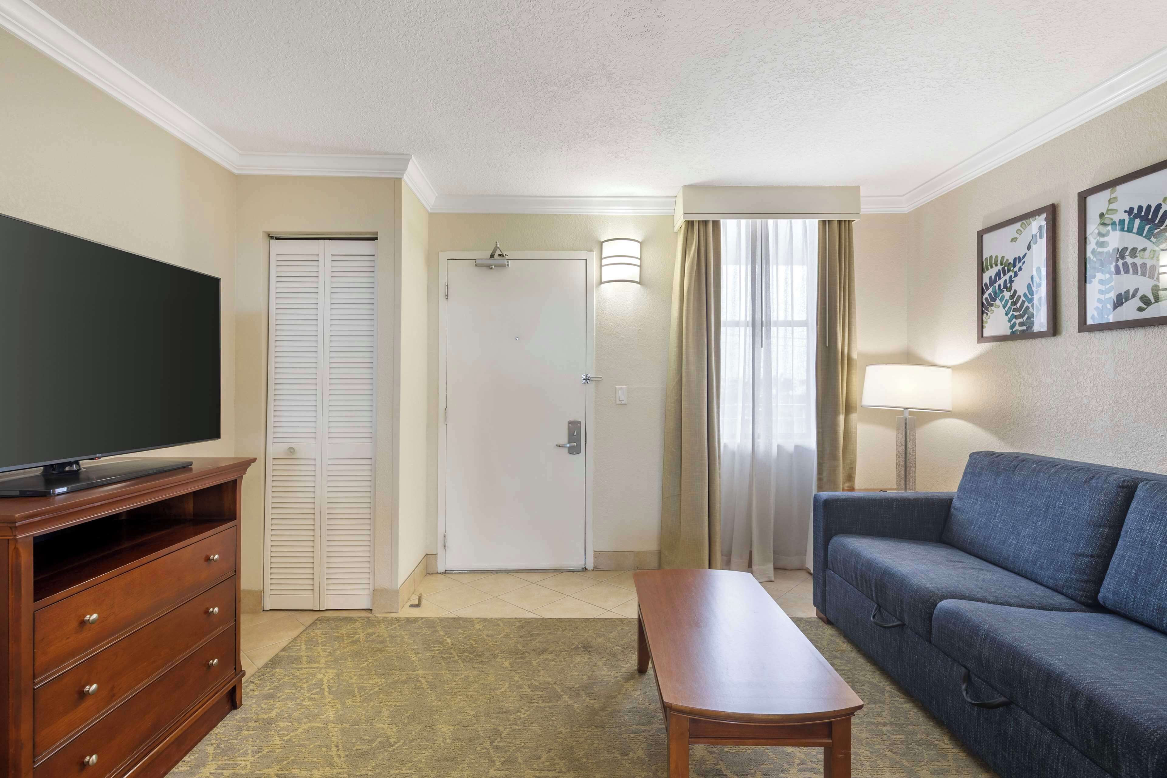 A hotel room with a wooden dresser, a flat-screen TV, a sofa, a coffee table, and a door leading to a bathroom.