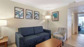 Una sala de estar con un sofá azul, un sillón estampado, una mesa de centro de madera y una lámpara de pie. Hay tres cuadros con motivos de hojas en la pared.