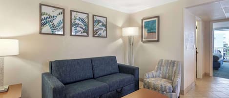 A living room with a blue sofa, a patterned armchair, a wooden coffee table, and a floor lamp. There are three framed leaf artworks on the wall.