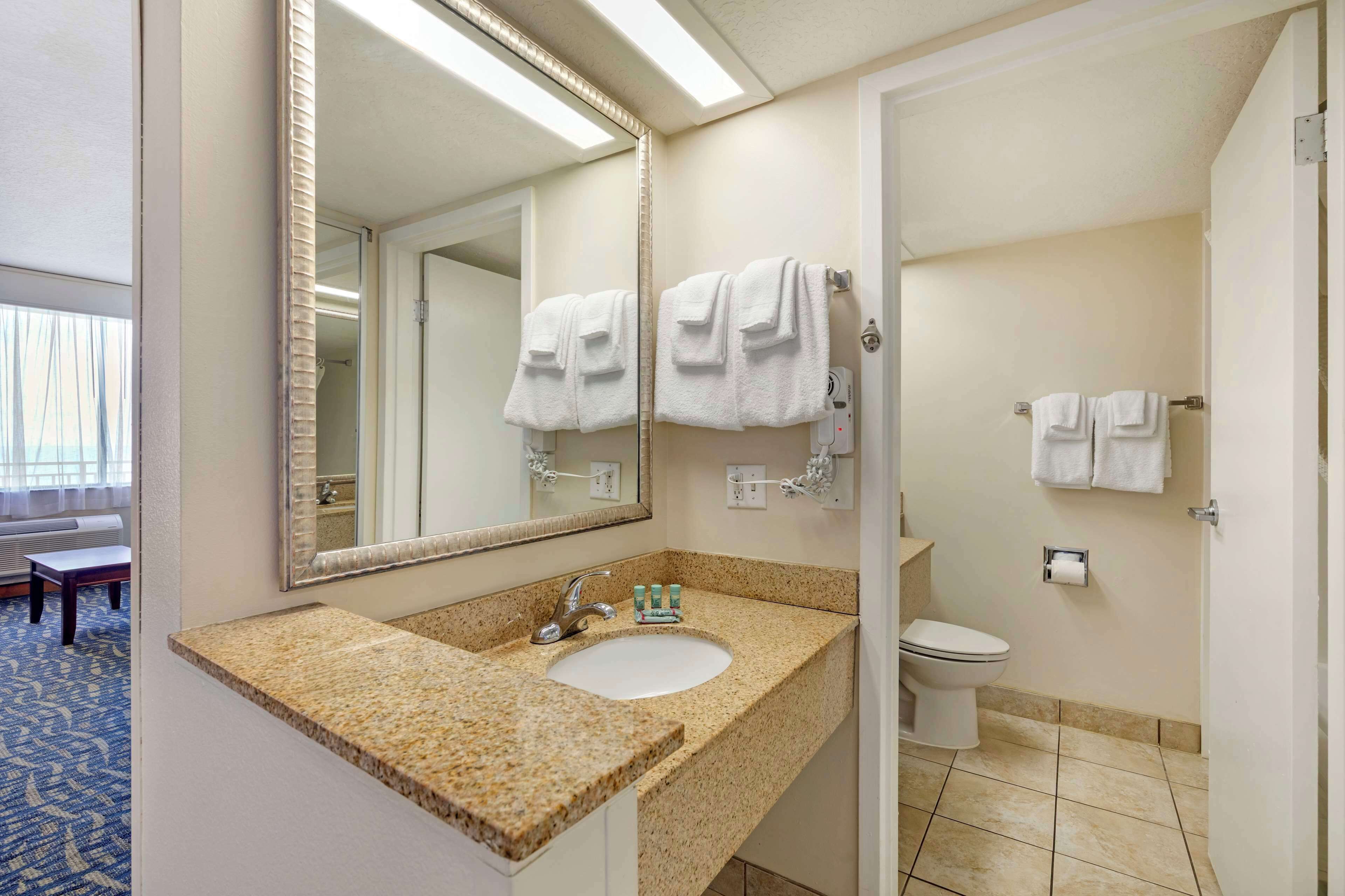 A hotel bathroom with a granite countertop sink, a large mirror, a toilet, and a towel rack.