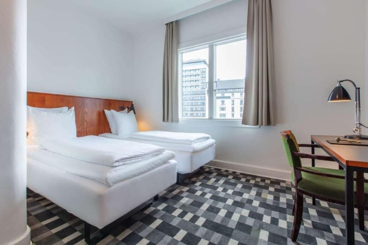 Une chambre d’hôtel avec deux lits, un bureau, une chaise, une fenêtre avec des rideaux et un sol à carreaux.