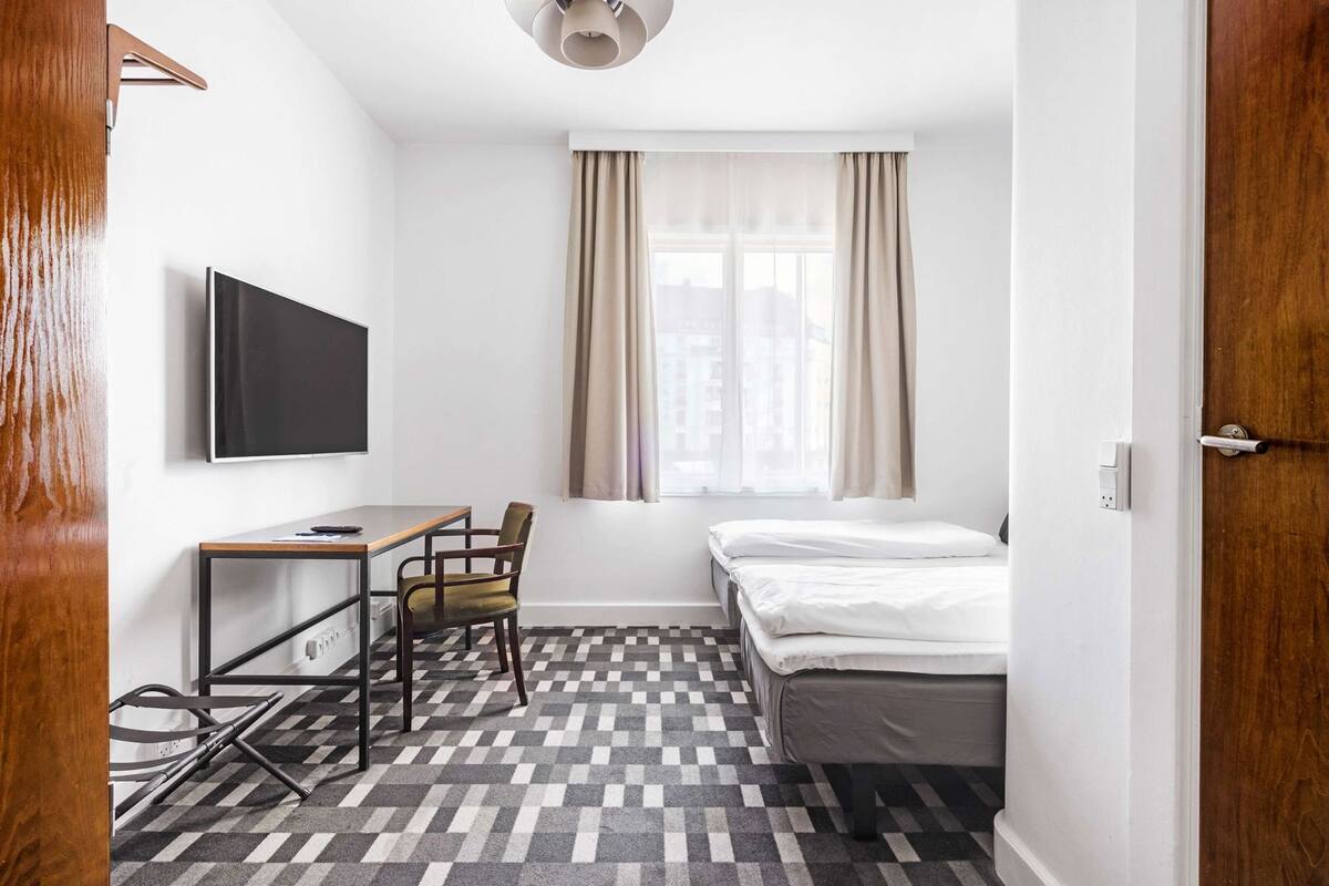 A small, tidy room with a bed, a desk, a chair, a TV, and a window with curtains.