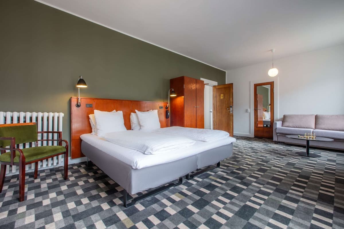 A hotel room with a large bed, a wooden headboard, a green chair, a sofa, and a patterned floor.