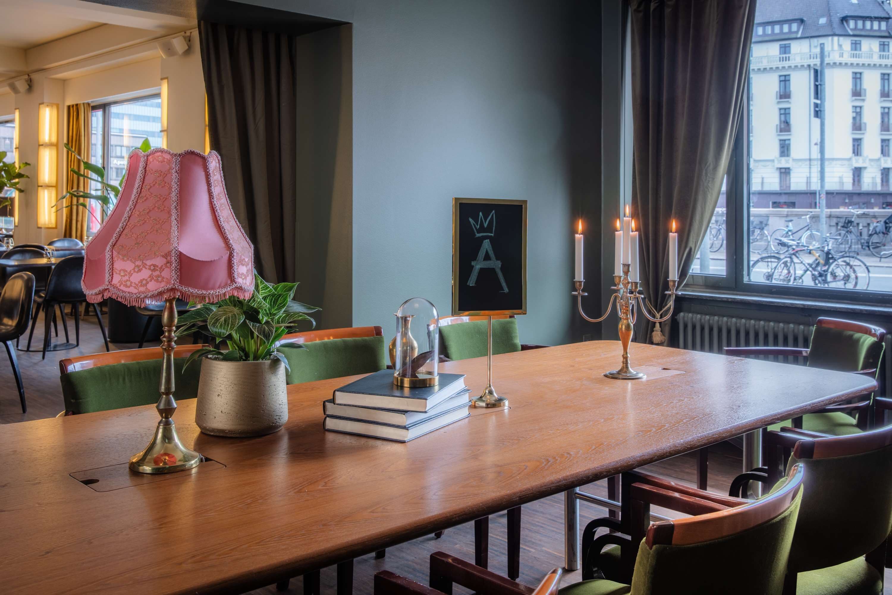 Un comedor de hotel con una larga mesa de madera, sillas tapizadas de color verde, una planta en maceta, una lámpara decorativa, una obra de arte enmarcada y un candelabro con velas.