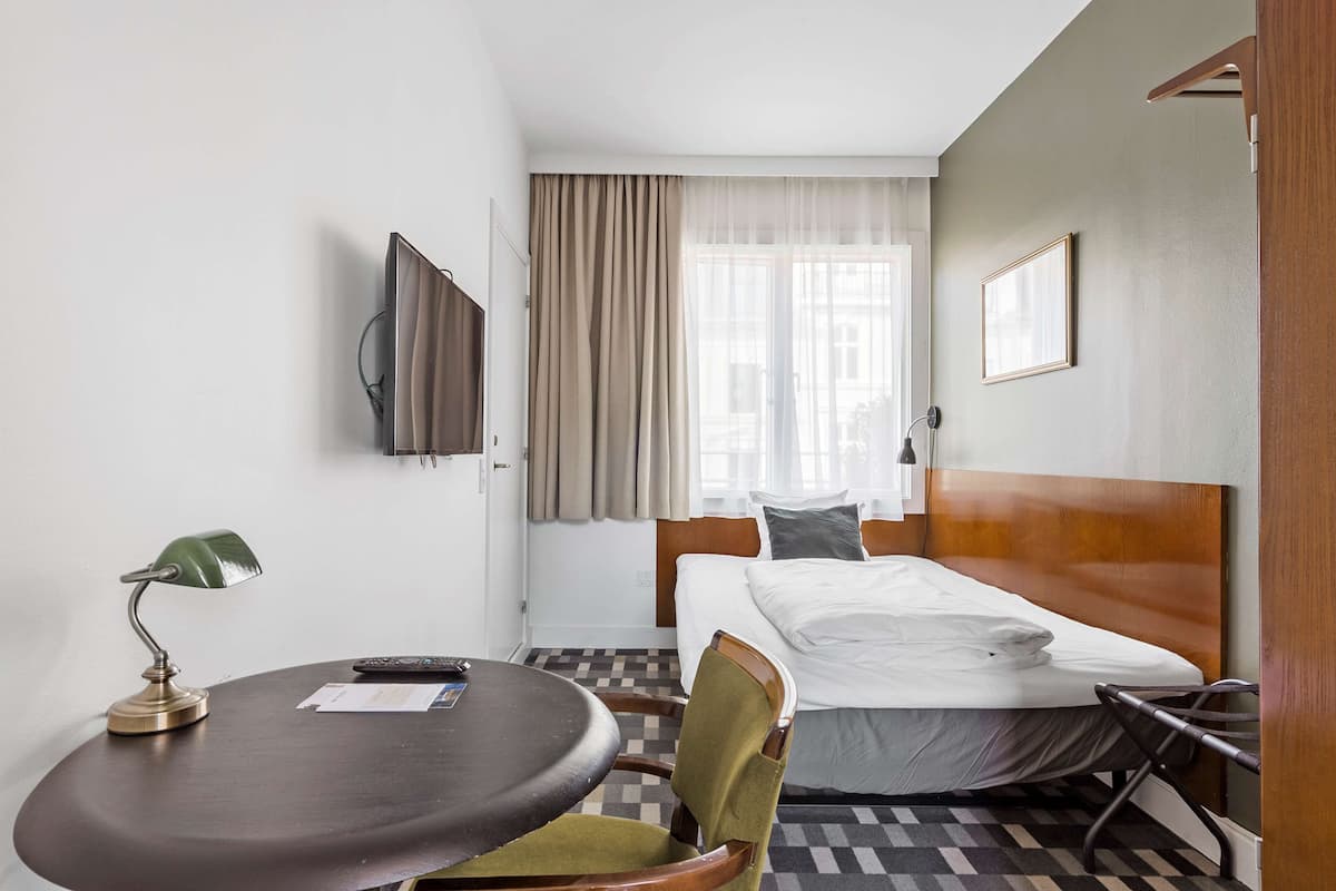 A hotel room with a bed, a desk with a lamp, a chair, a television, and a window with curtains.