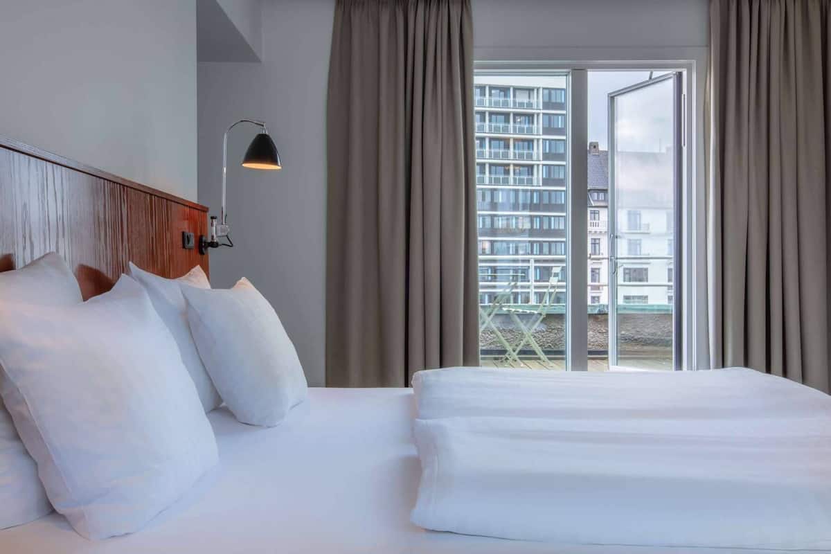 A hotel room with a large bed, a desk, a lamp, and a view of buildings through the window.
