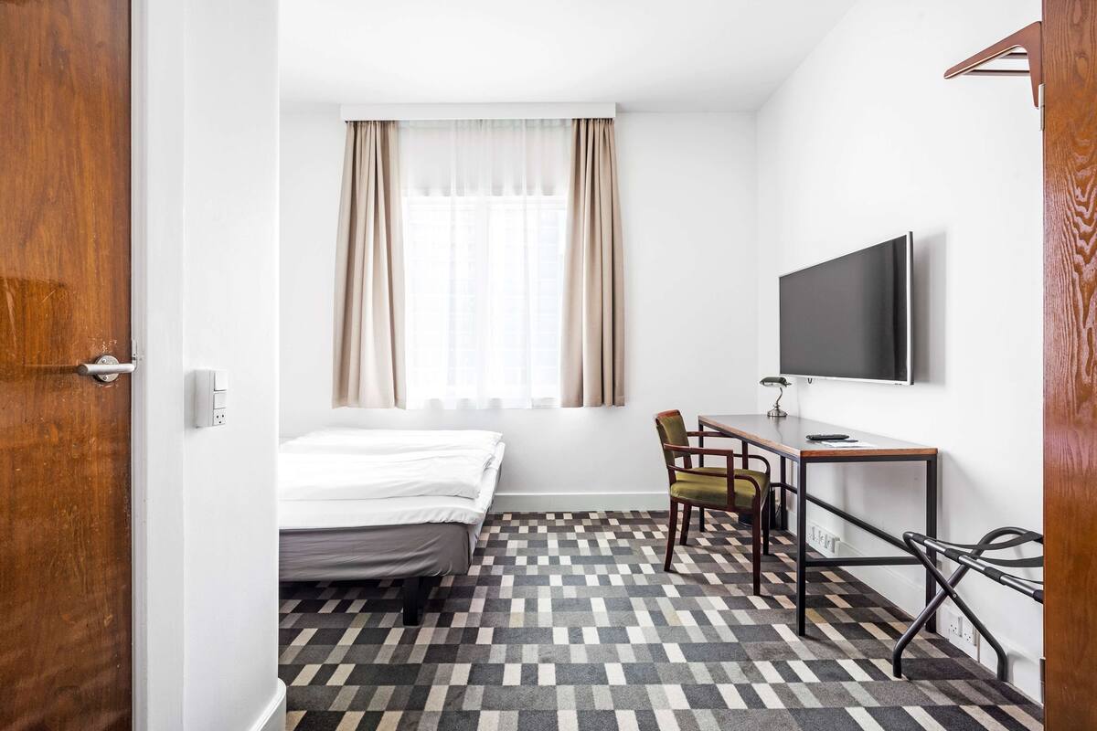 A hotel room with a bed, a desk with a chair, a television, and a patterned floor.