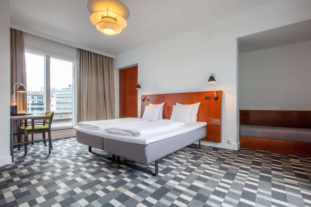 A hotel room with a large bed, a desk, a chair, a window with curtains, and a patterned floor.