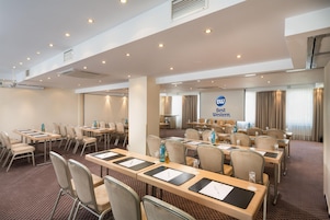 Un salón de conferencias con varias mesas y sillas, conductos de aire acondicionado y una pantalla grande que muestra el logotipo 'Best Western'.