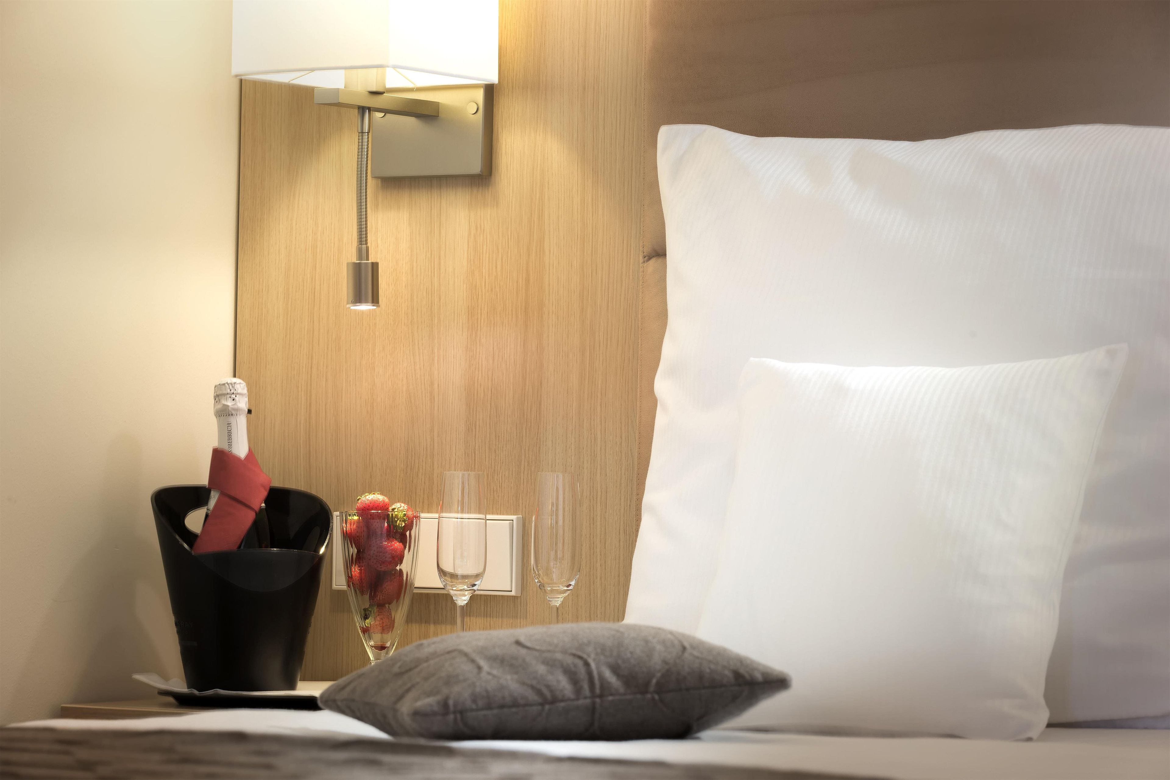 A hotel room with a bed, a nightstand, a bottle of champagne, two glasses, and a vase with strawberries.