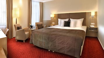 A hotel room with a large bed, two armchairs, a small table with a bottle of champagne, and a nightstand with a lamp.
