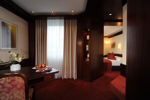 Hotelkamer met een bed, een bureau met een bloevas, een rood tapijt en uitzicht op een andere kamer.