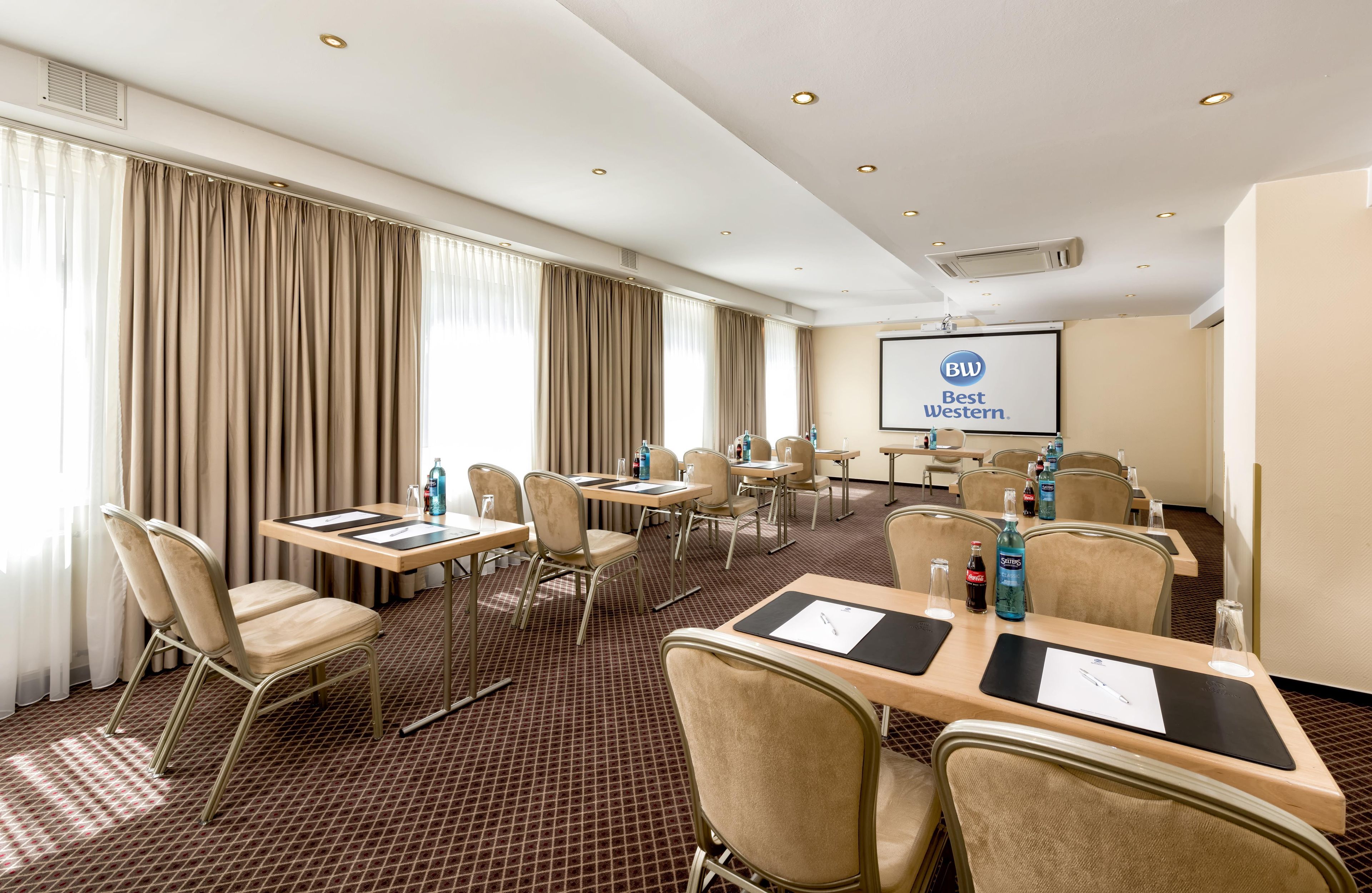 Ein Konferenzraum mit Tischen, Stühlen, einer Projektionsleinwand mit dem Schriftzug 'Best Western' und einem großen Fenster mit Vorhängen.