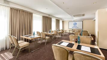 Ein Konferenzraum mit Tischen, Stühlen, einer Projektionsleinwand mit dem Schriftzug 'Best Western' und einem großen Fenster mit Vorhängen.