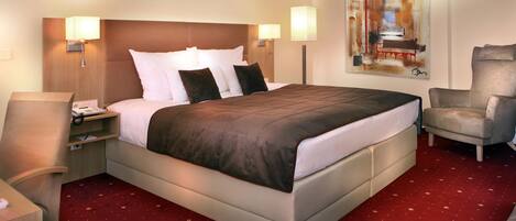 A hotel room with a large bed, two bedside lamps, a chair, a desk with a phone, and a painting on the wall.