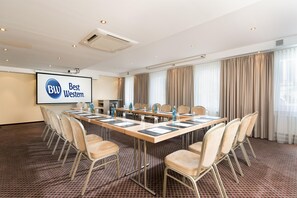 Ein Konferenzraum mit einem langen Tisch, Stühlen, einer Projektionsleinwand und einem großen Fenster mit Vorhängen.