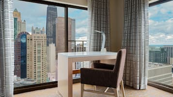 A hotel room with a large window offering a city view, a desk with a chair, and a modern lamp.