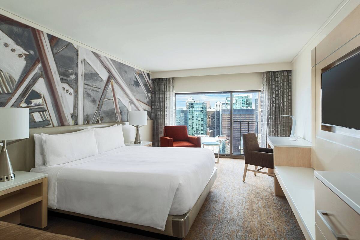 Habitación de hotel con una cama grande, un escritorio, una silla, un televisor y vistas a la ciudad desde la ventana.
