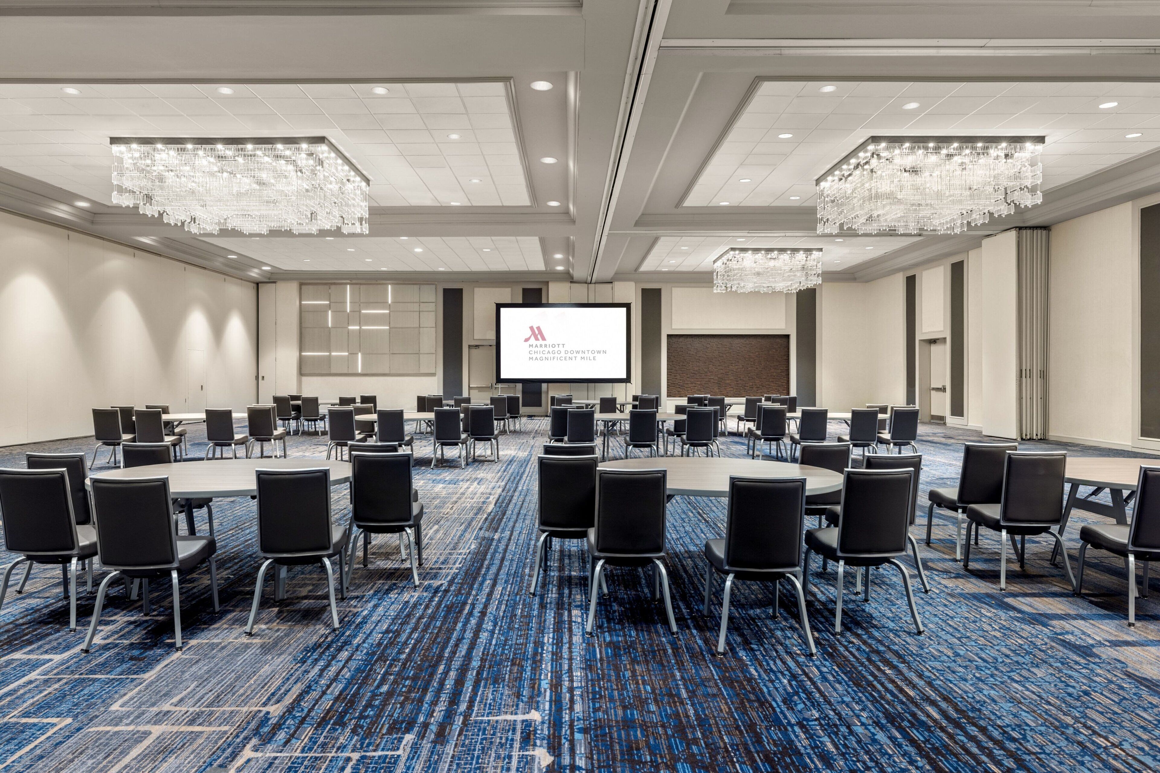 A conference room with a large screen, multiple tables, and chairs, featuring a blue carpet and chandeliers.