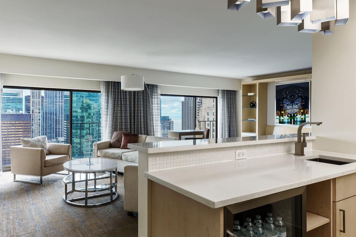 Habitación de hotel moderna con un ventanal que ofrece vistas a la ciudad, una zona de cocina con encimera blanca y un espacio de descanso con una mesa de centro redonda.