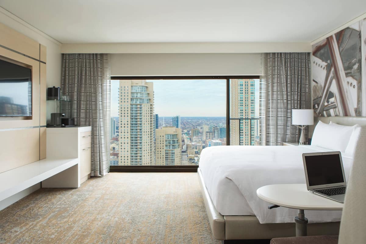 Habitación de hotel con una cama grande, un escritorio con una computadora portátil, un televisor y vistas a edificios altos.