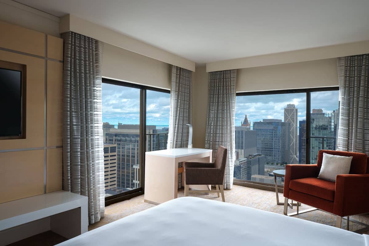 Habitación de hotel con una cama grande, un escritorio, una silla y un televisor. Amplias ventanas con cortinas y vistas a la ciudad.