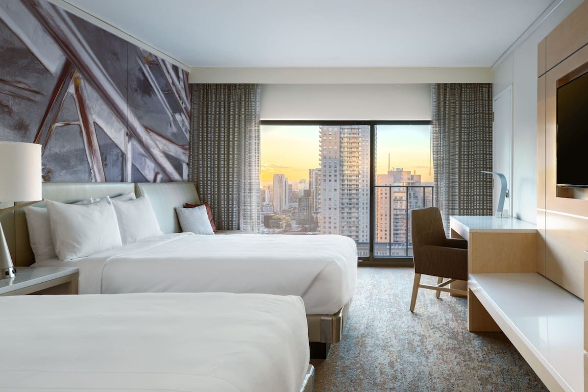 Habitación de hotel con dos camas, un escritorio, una silla y un ventanal con vistas a la ciudad.