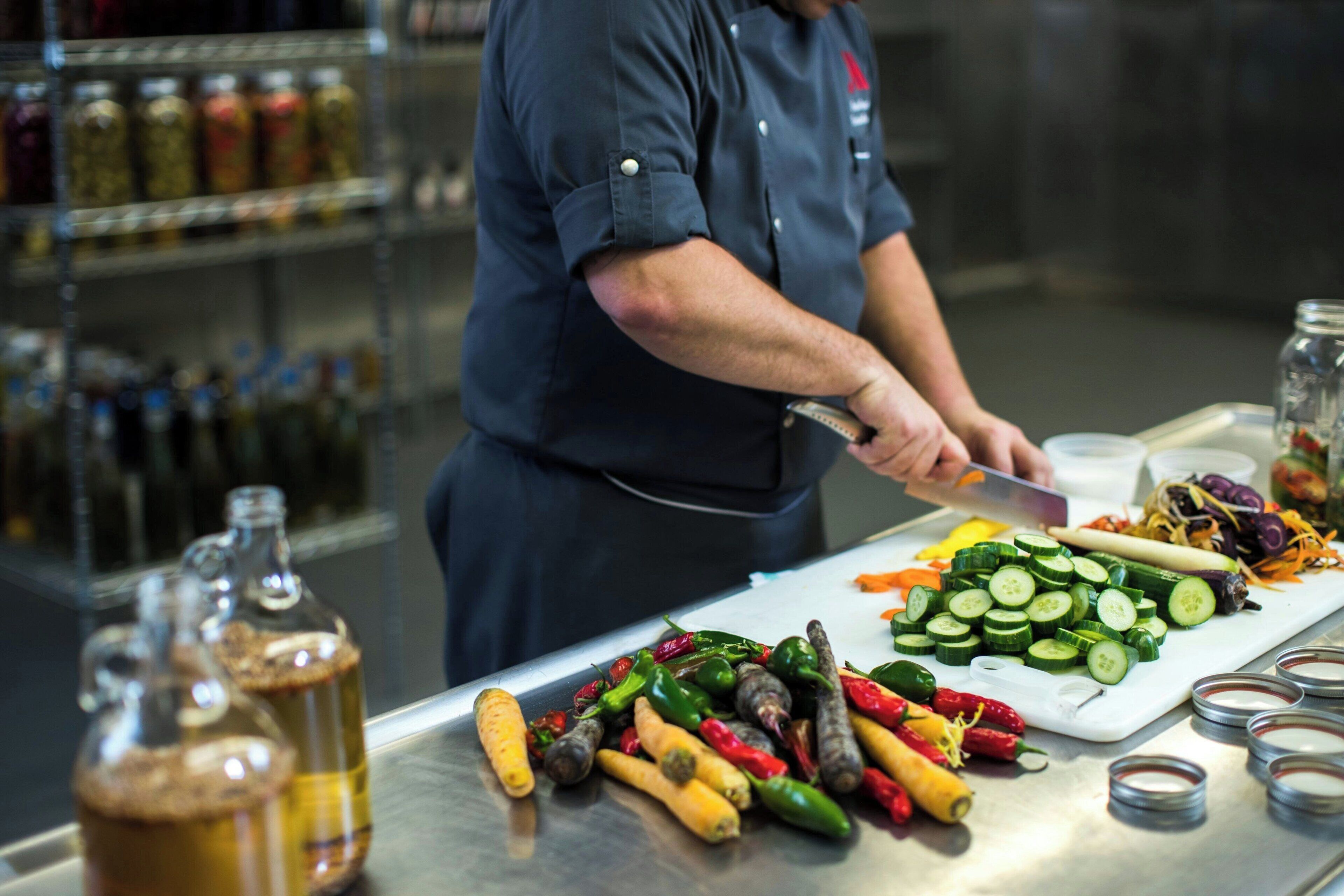 Un chef prépare des plats à base de légumes et de sauces sur un plan de travail en acier inoxydable.