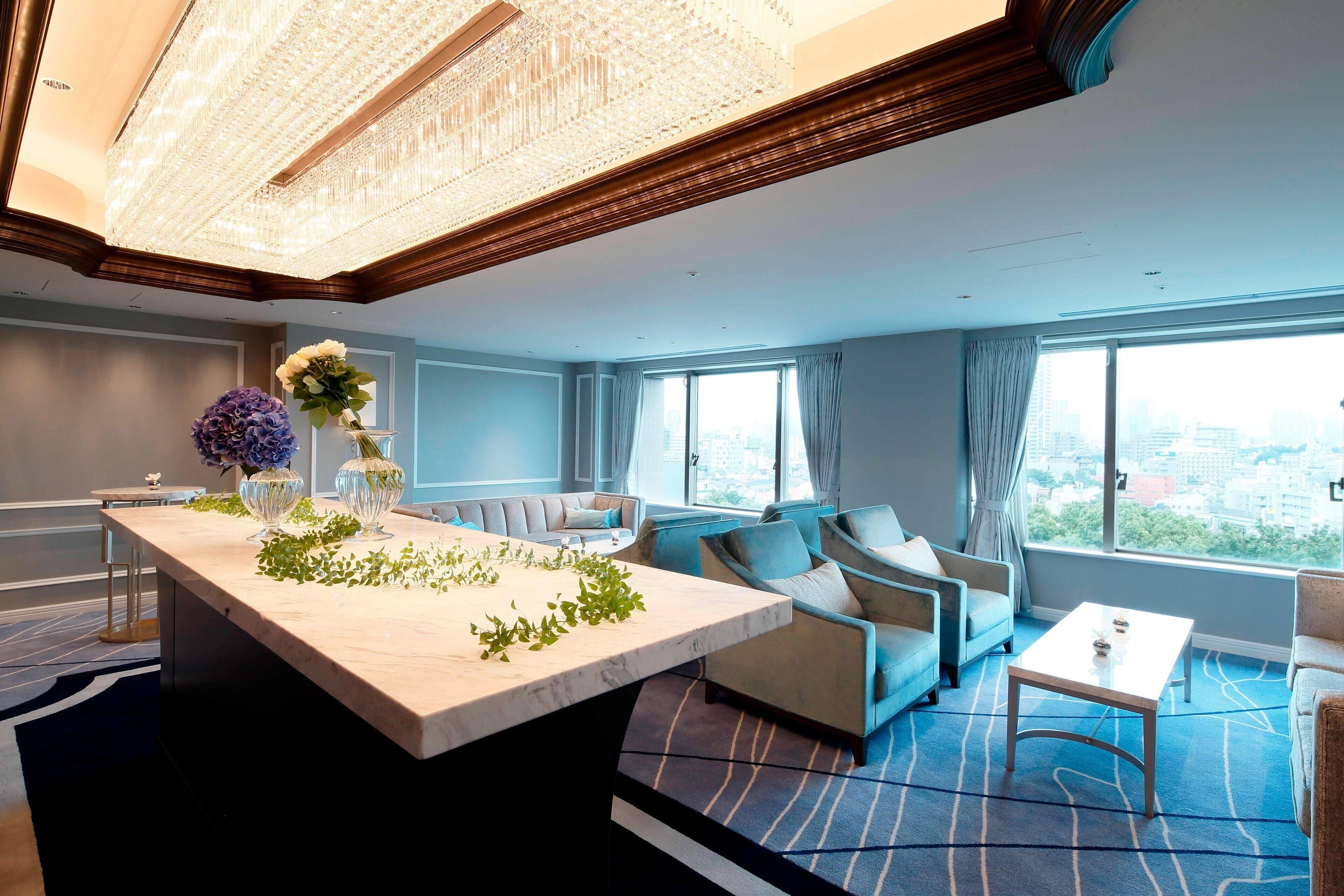 A modern hotel room with a marble island, a chandelier, a sofa, a coffee table, and large windows with a city view.