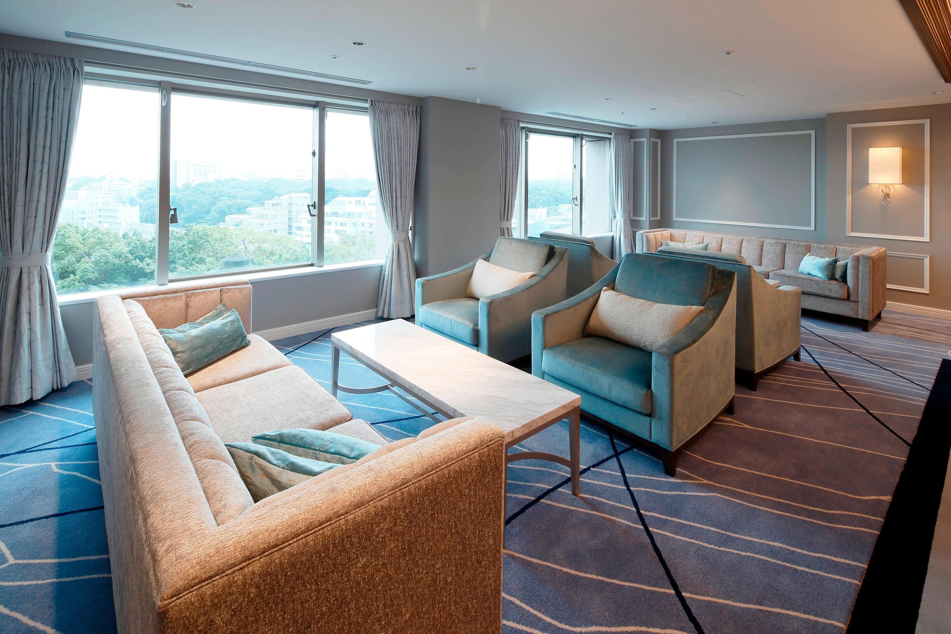 A modern hotel room with a large window, a sofa, two armchairs, a coffee table, and a patterned carpet.