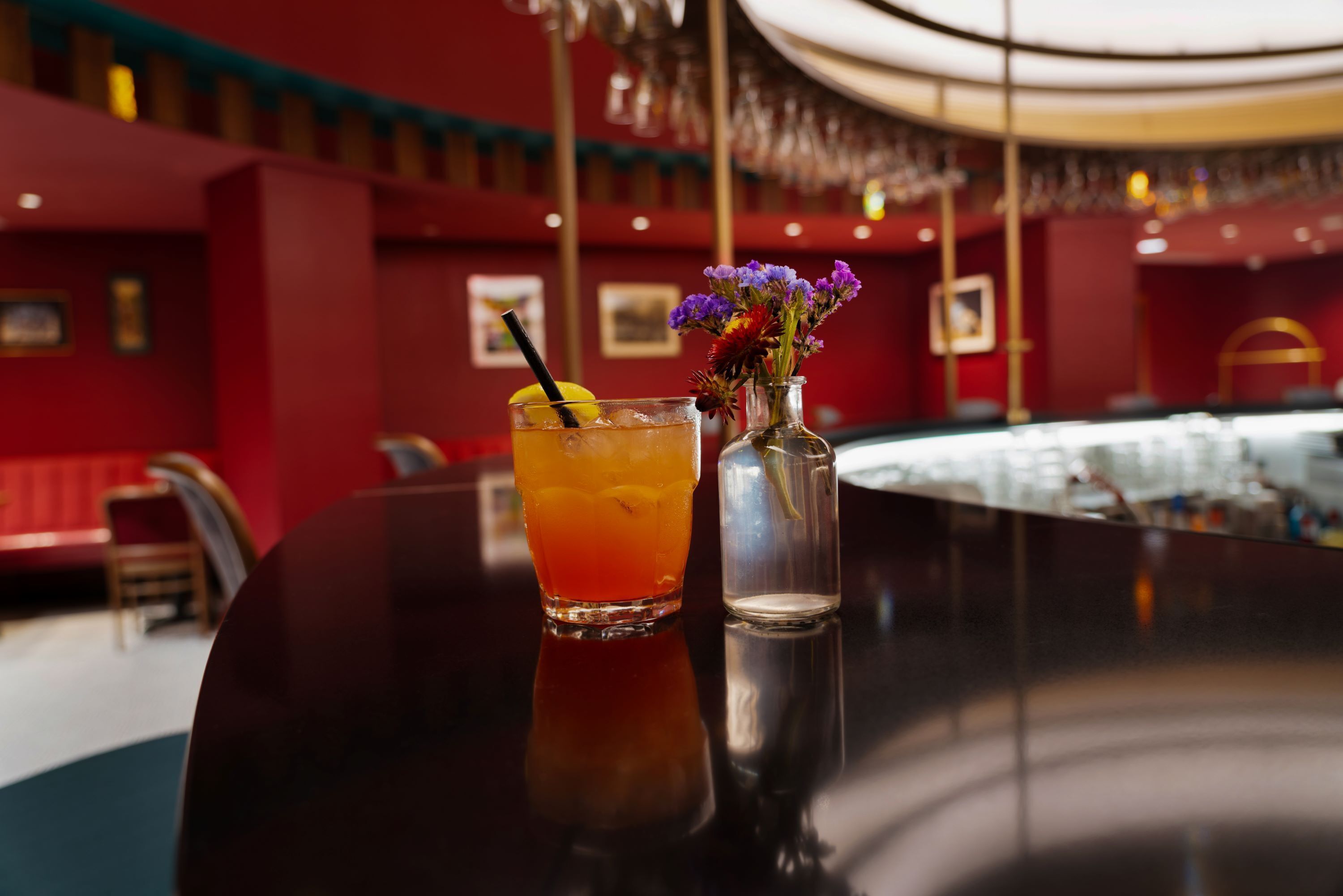 A bar area with a glass of amber-colored drink, a straw, and a small vase with purple flowers.