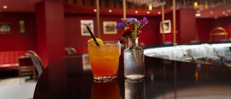 A bar area with a glass of amber-colored drink, a straw, and a small vase with purple flowers.
