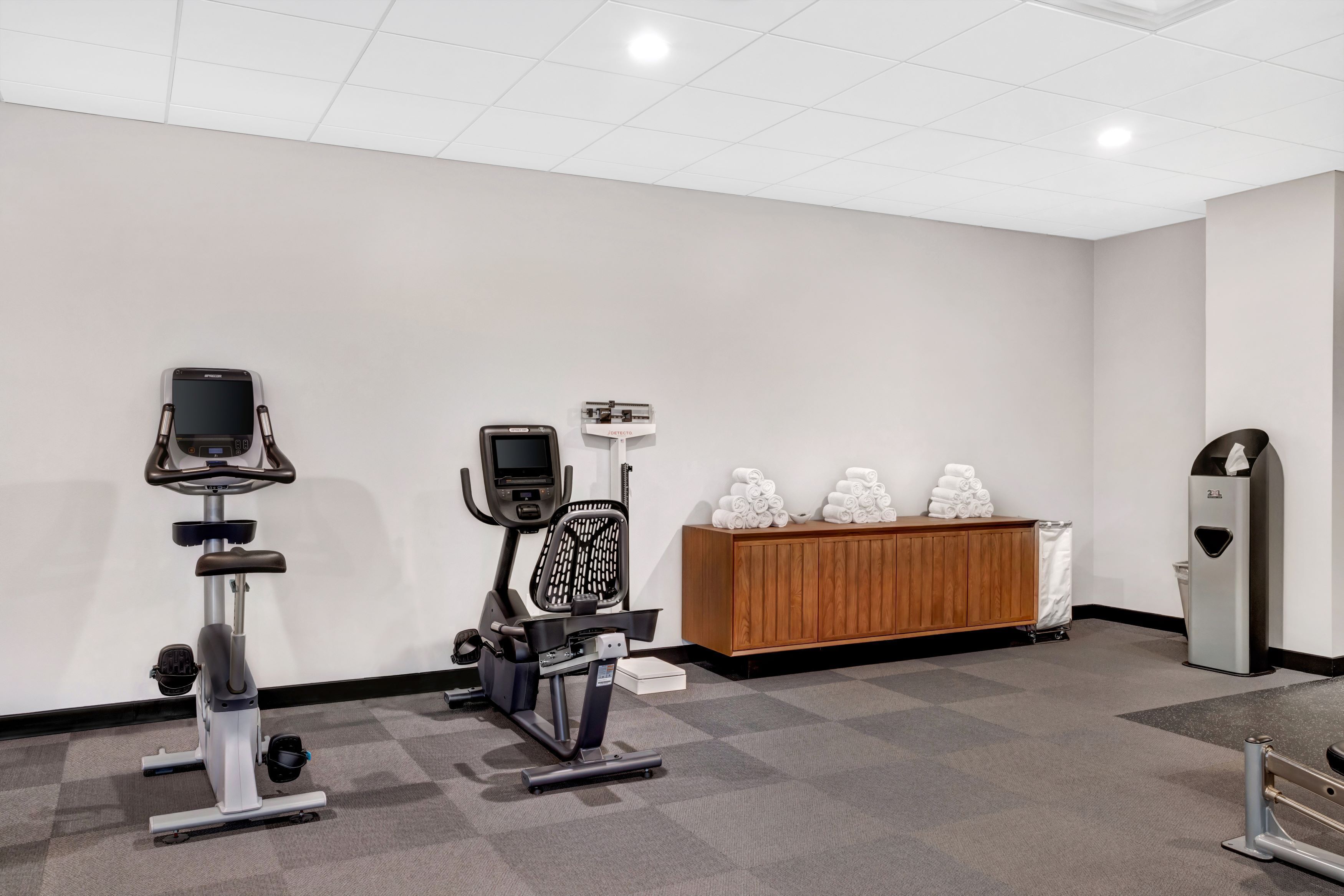 A fitness room with exercise bikes, a wooden cabinet with towels, and a trash bin.