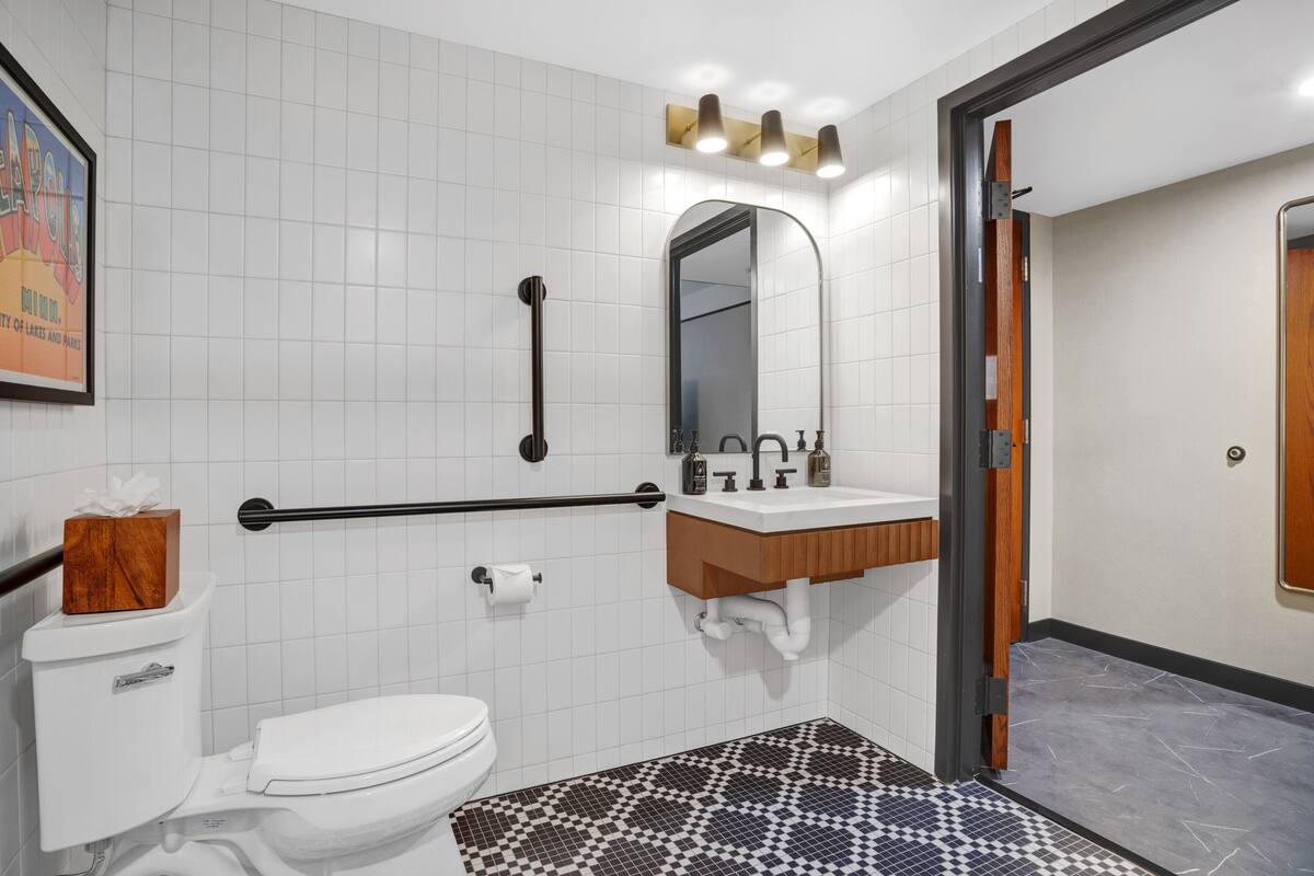 Ein modernes Badezimmer mit weißen Fliesen, einem Holzkosmetikschrank, einer schwarzen Haltestange, einer weißen Toilette und einem gemusterten Boden.