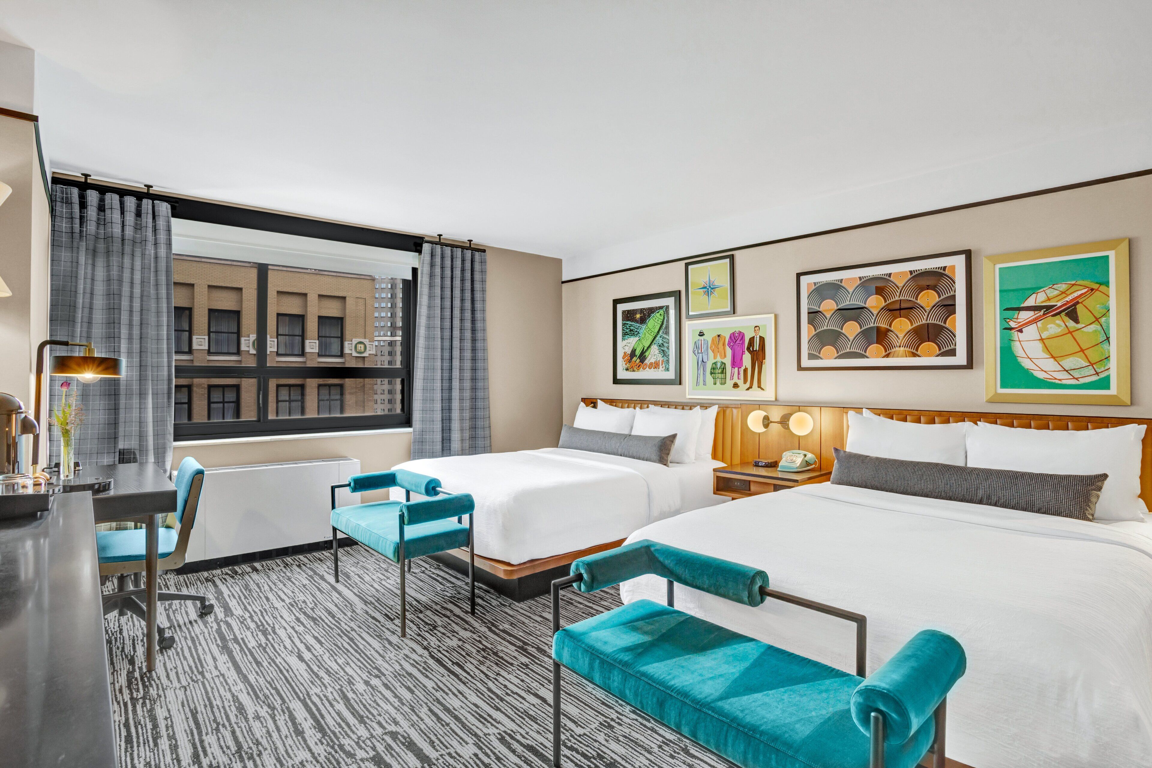 A hotel room with two beds, a desk, a chair, and a view of buildings through a window.
