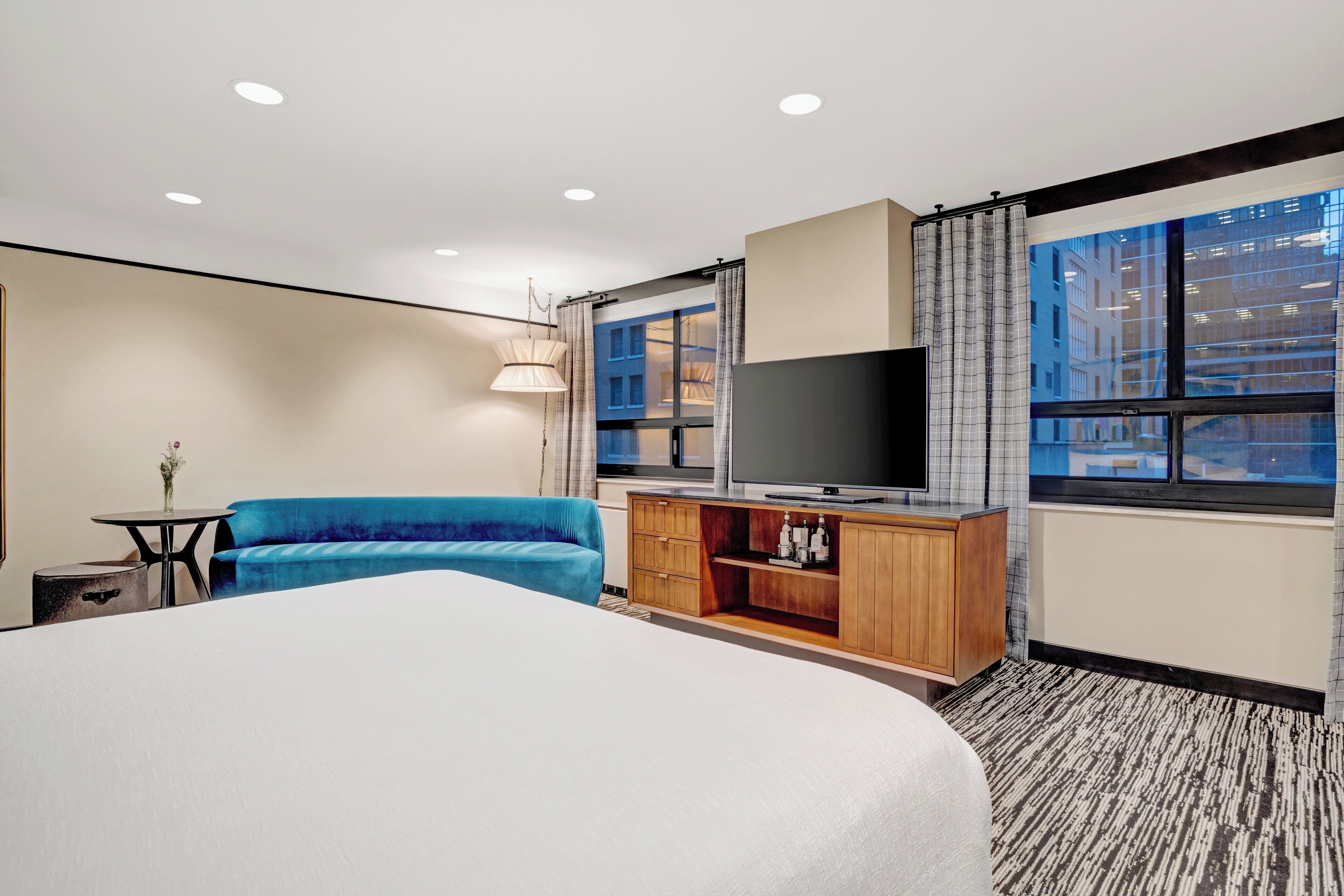 A hotel room with a large bed, a blue sofa, a television on a wooden cabinet, and a window with a city view.