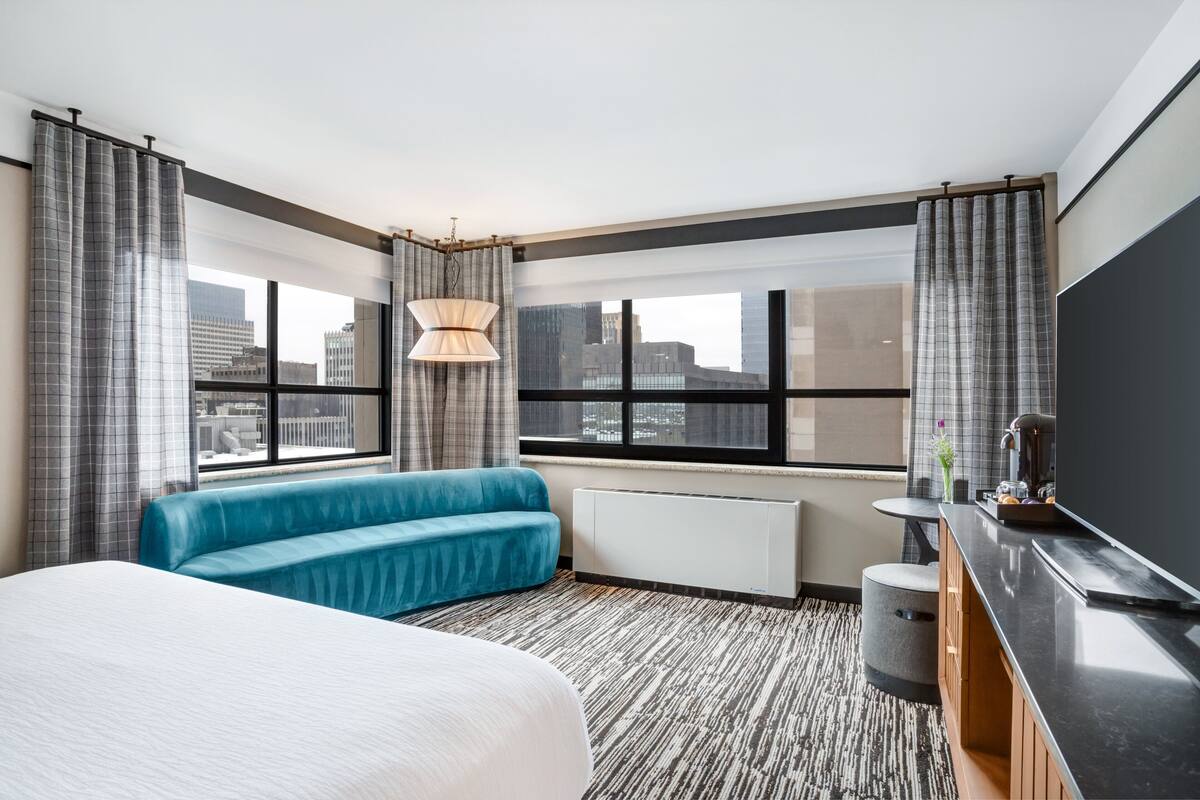 Ein Hotelzimmer mit einem großen Bett, einem blauen Sofa, einem kleinen Tisch, einem Fernseher und einem Fenster mit Blick auf die Stadt.