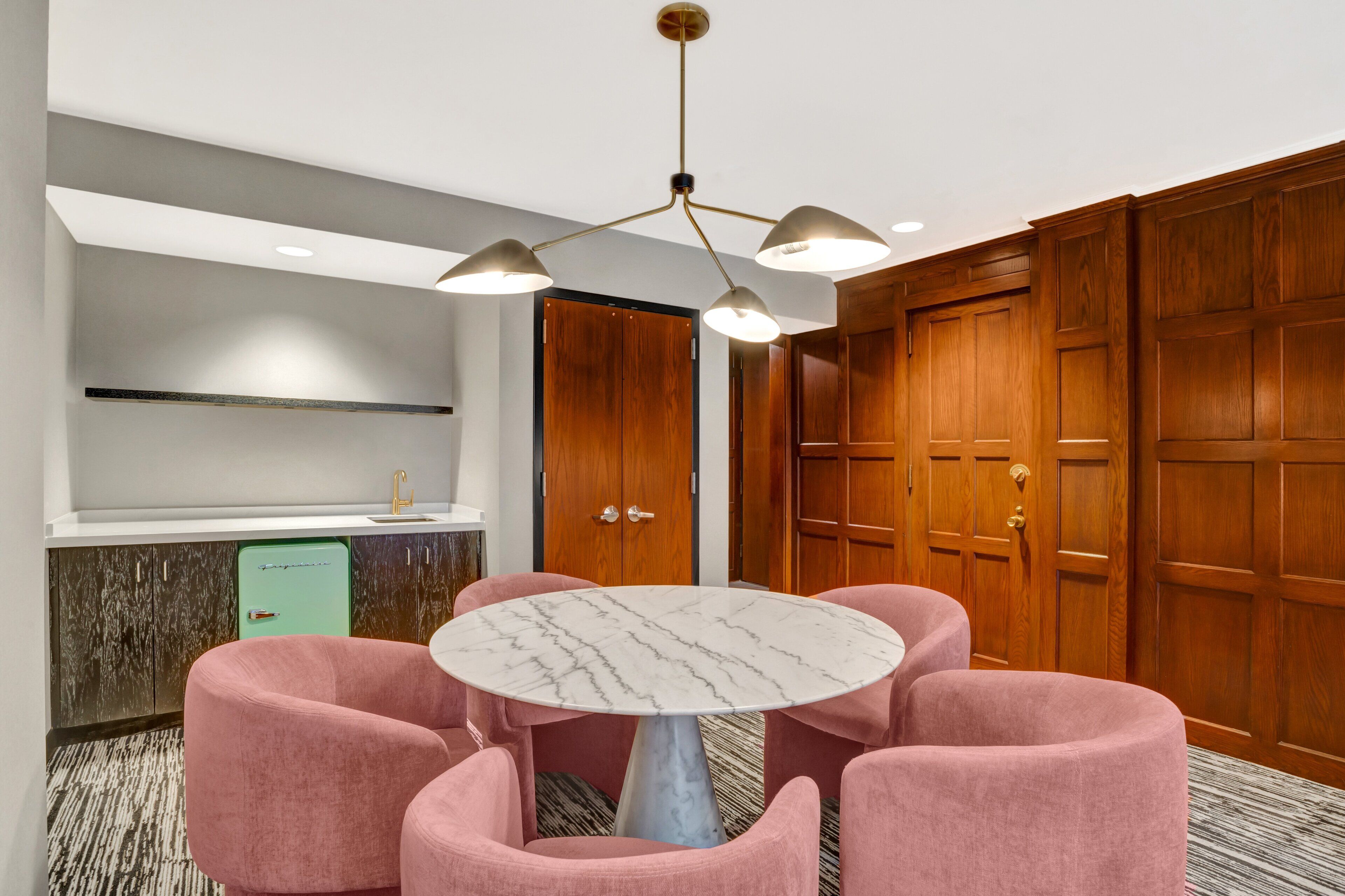 Una habitación de hotel moderna con una mesa de mármol rodeada de sillas rosadas, puertas de madera y una kitchenette con un refrigerador verde.