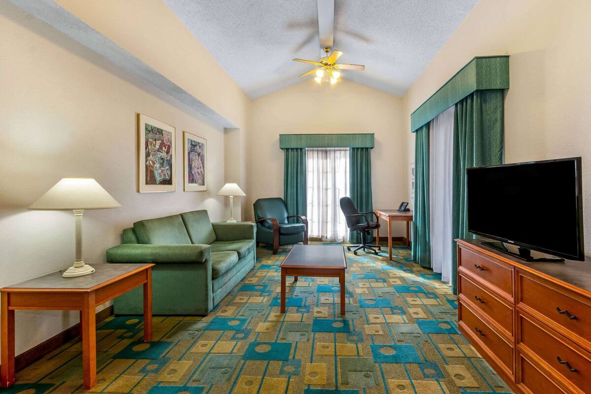 A hotel room with a green sofa, a wooden table, a TV on a wooden cabinet, and a patterned carpet.