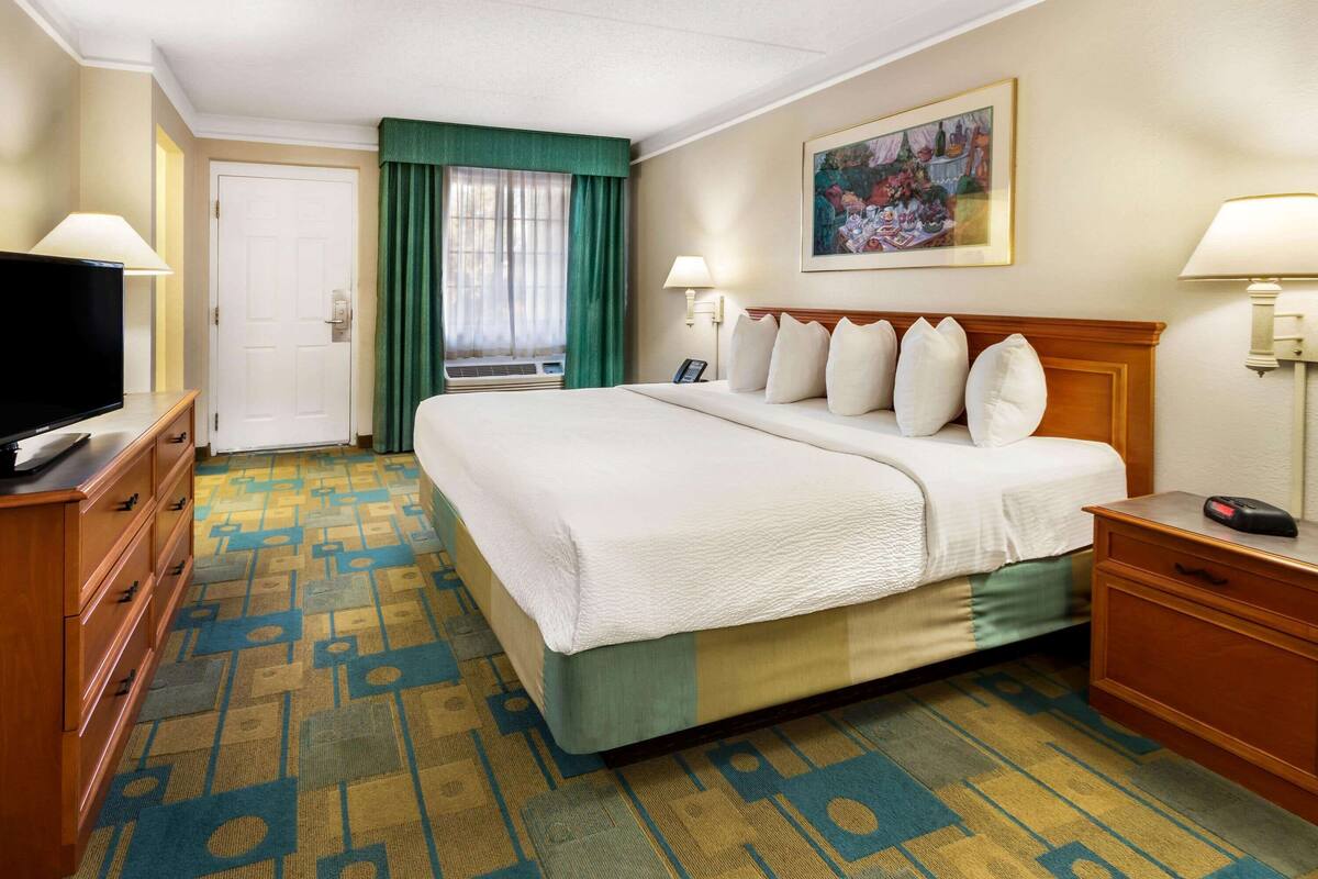 A hotel room with a large bed, a television on a dresser, a desk with a lamp, and a painting on the wall.
