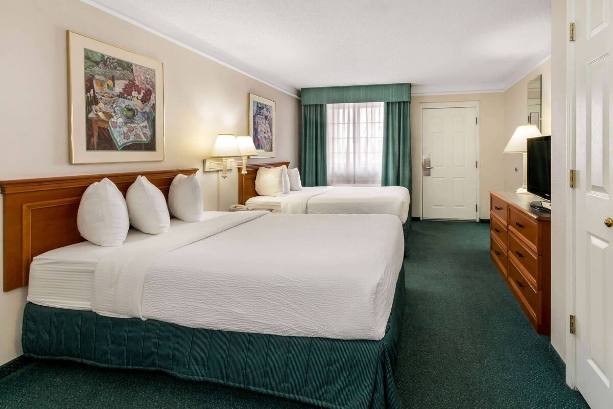 A hotel room with two beds, a wooden headboard, a desk with a lamp, a television, and artwork on the walls.