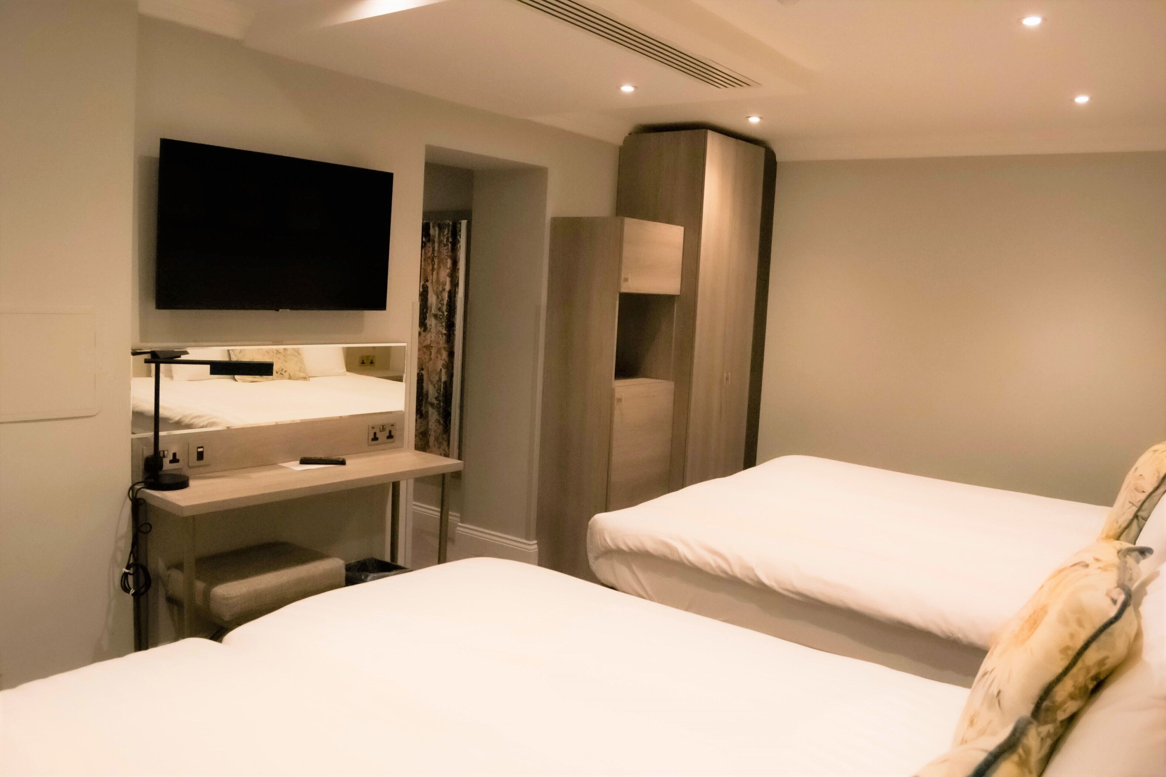 A hotel room with two beds, a flat-screen TV mounted on the wall, a small desk with a lamp, and a built-in wardrobe.