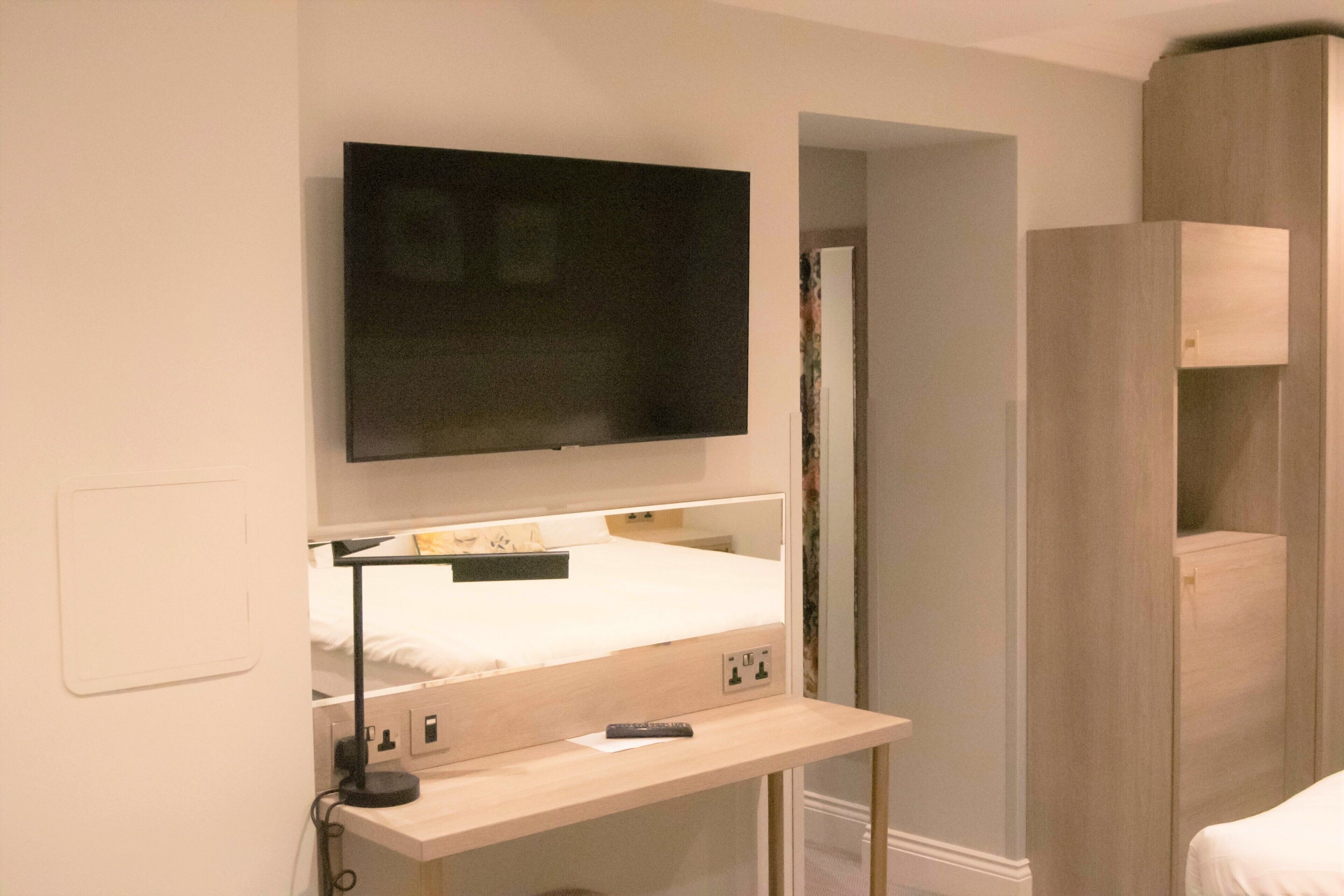 A bedroom with a bed, a TV mounted above it, a bedside table with a lamp, and a wooden wardrobe.